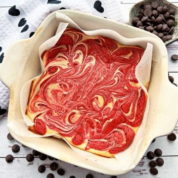 a pan of vibrant red and cream colored brownies on a white farm board with a black and white towel accent and bittersweet chocolate chips scatter around the baking dish