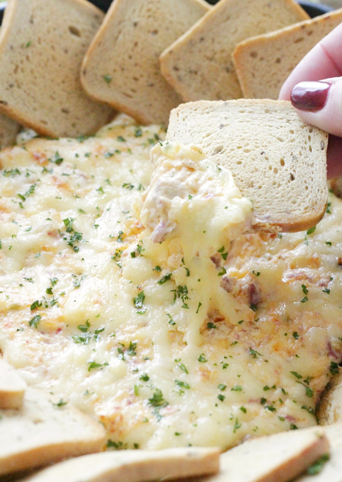 hand dipping rye bread square into ultimate reuben dip showing the melted cheese pull