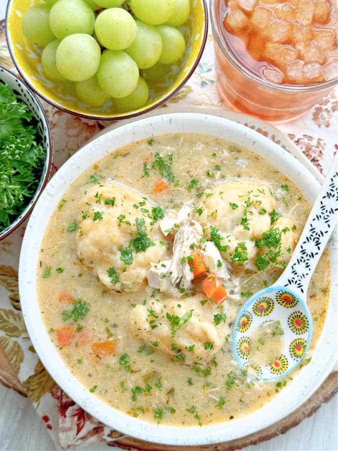 a bowl full of rotisserie chicken and dumplings pictured with a glass or iced tea and a bowl of green grapes