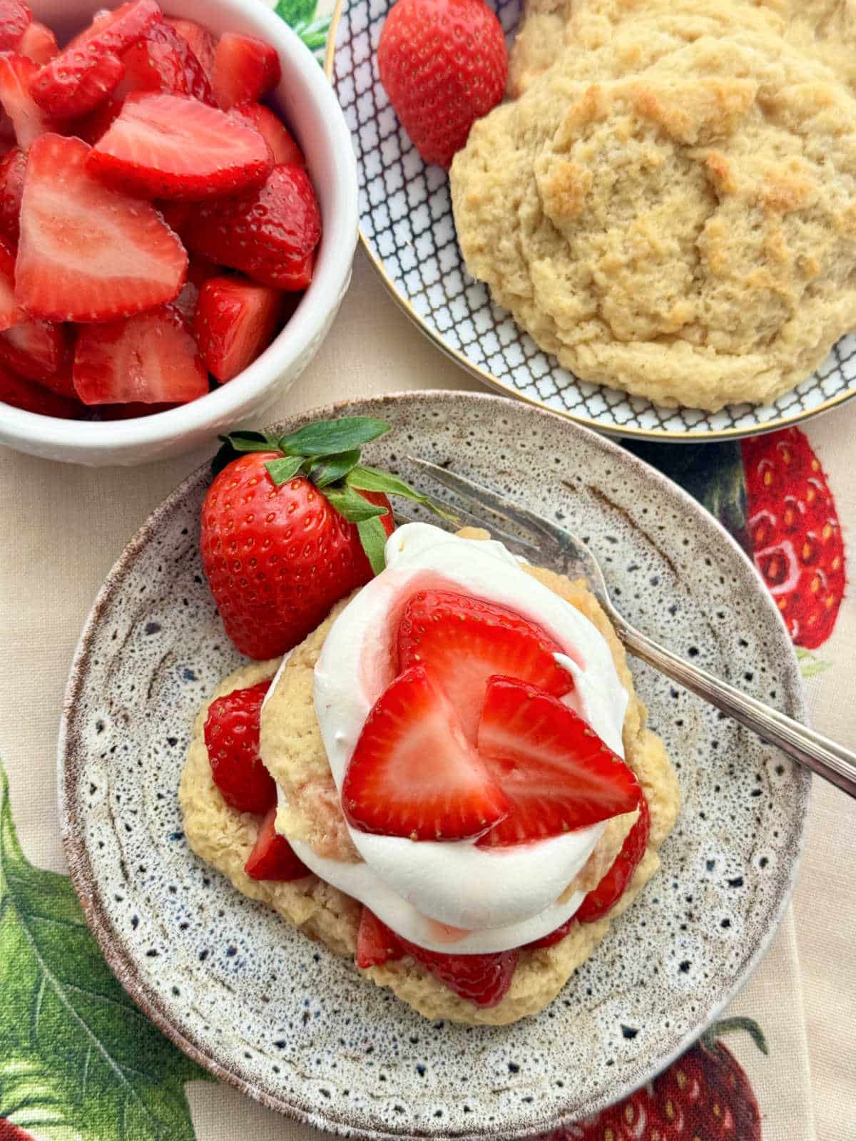 plated strawberry shortcake flanked by a plate of plain shortcakes and a bowl of juicy sliced strawberries