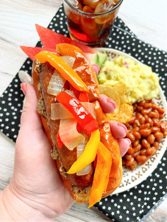 holding a loaded air fryer sausage in hand - in a bun, topped with peppers and onions and mustard