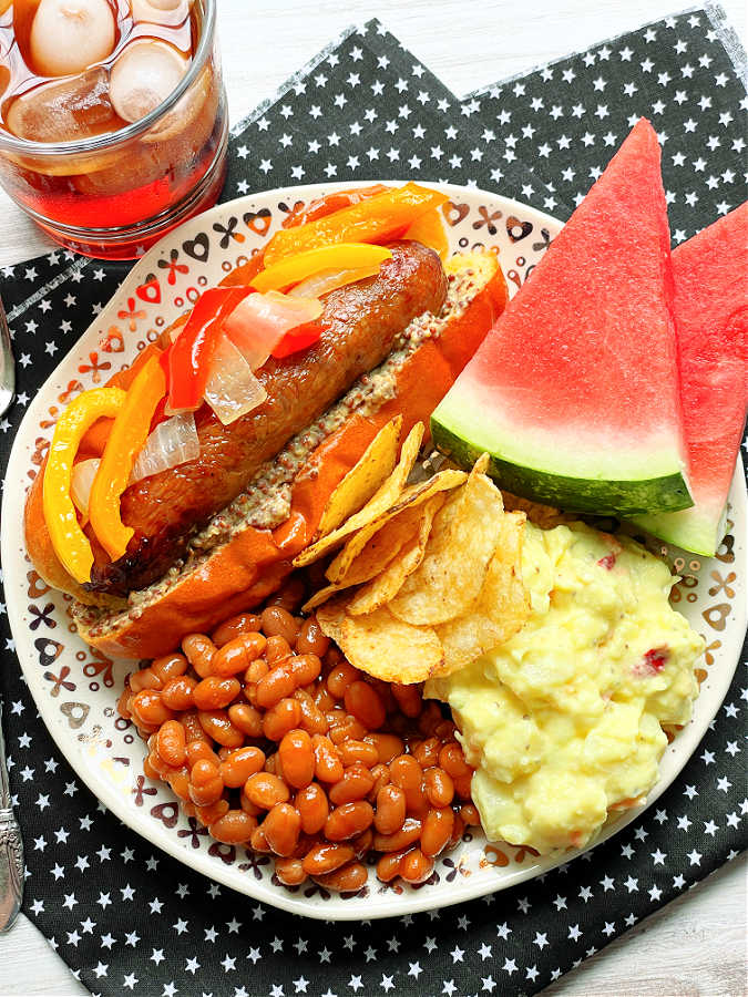 air fryer sausage in a bun with peppers plated with baked beans, potato salad, chips and watermelon