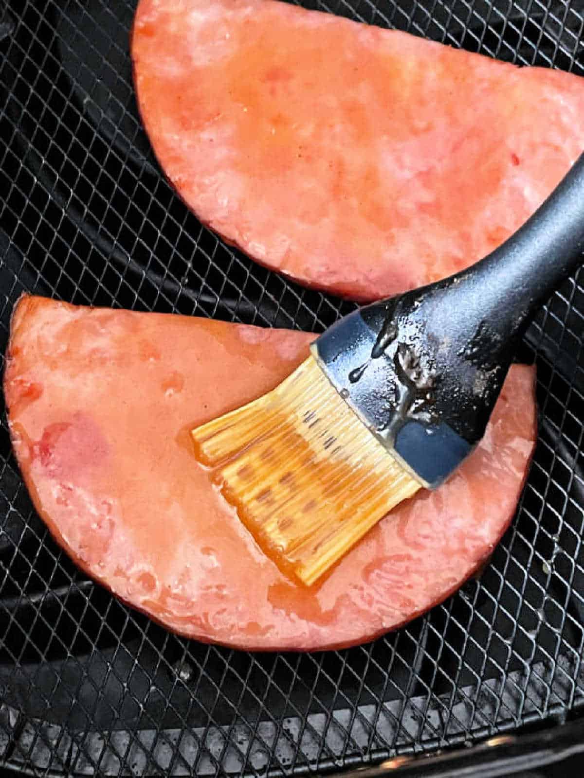ham steaks in the air fryer basket, being brushed with brown sugar glaze