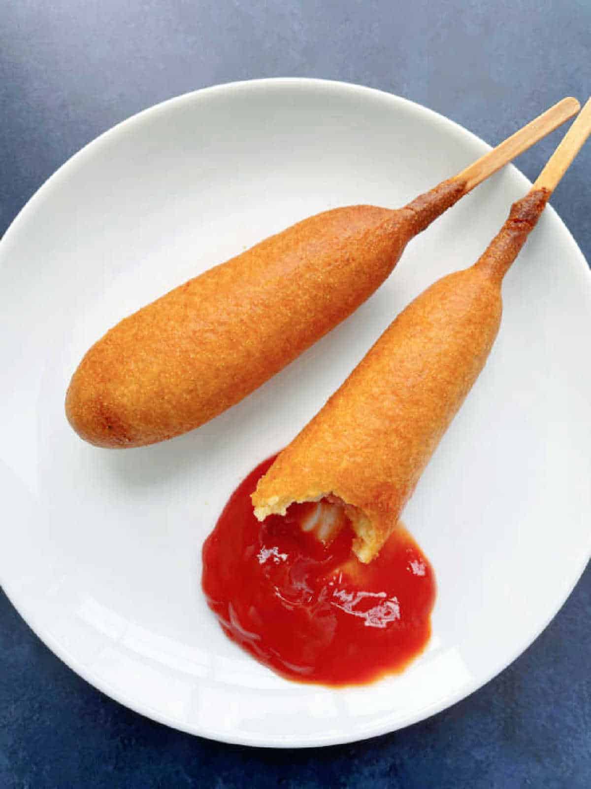 two corn dogs on a white plate with some ketchup for dipping