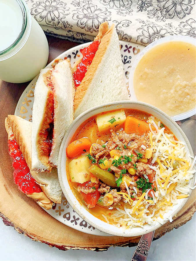 a bowl of campfire stew served with a peanut butter and jelly sandwich, applesauce and a glass of milk