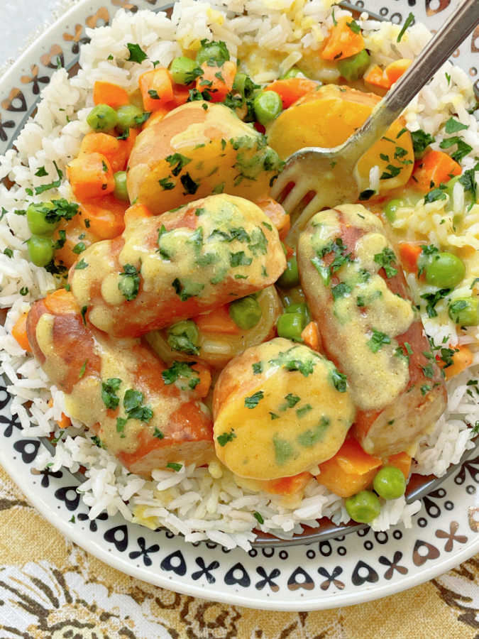 a plate full of rice topped with curried sausage, potatoes, peas and carrots