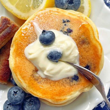 lemon blueberry pancakes topped with butter and syrup plated with sausage and fresh blueberries