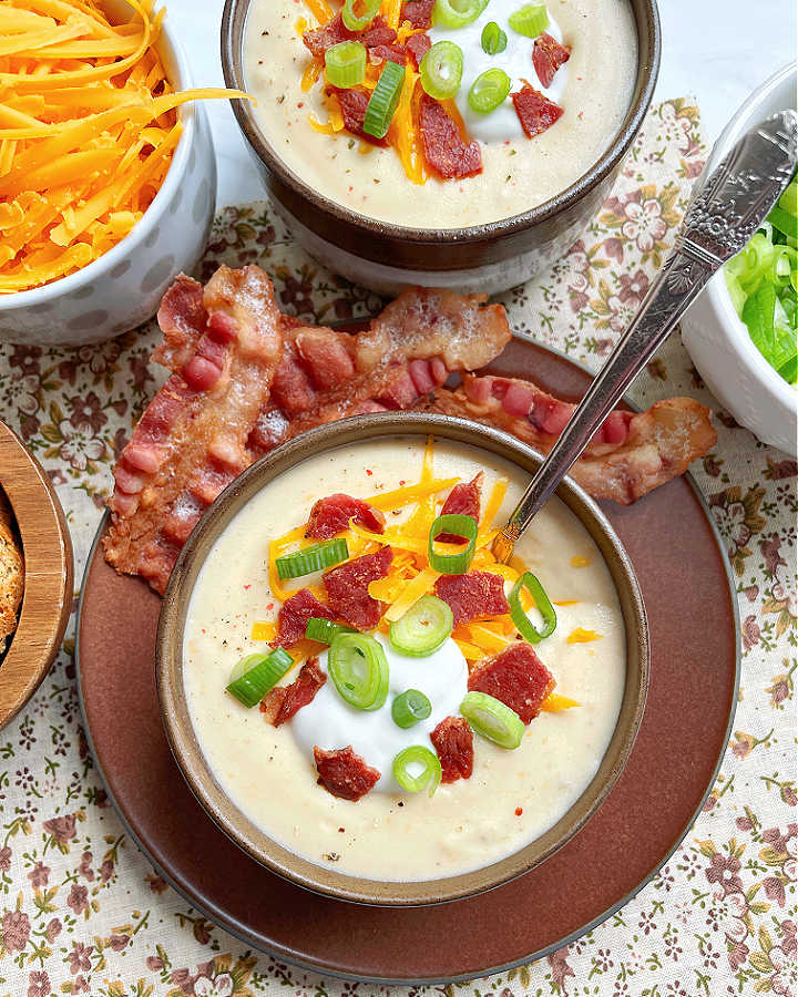 beautiful bowl of creamy crockpot potato soup garnished with cooked and crumbled bacon, green onions, sour cream and shredded cheese - with extra bacon slices on the side
