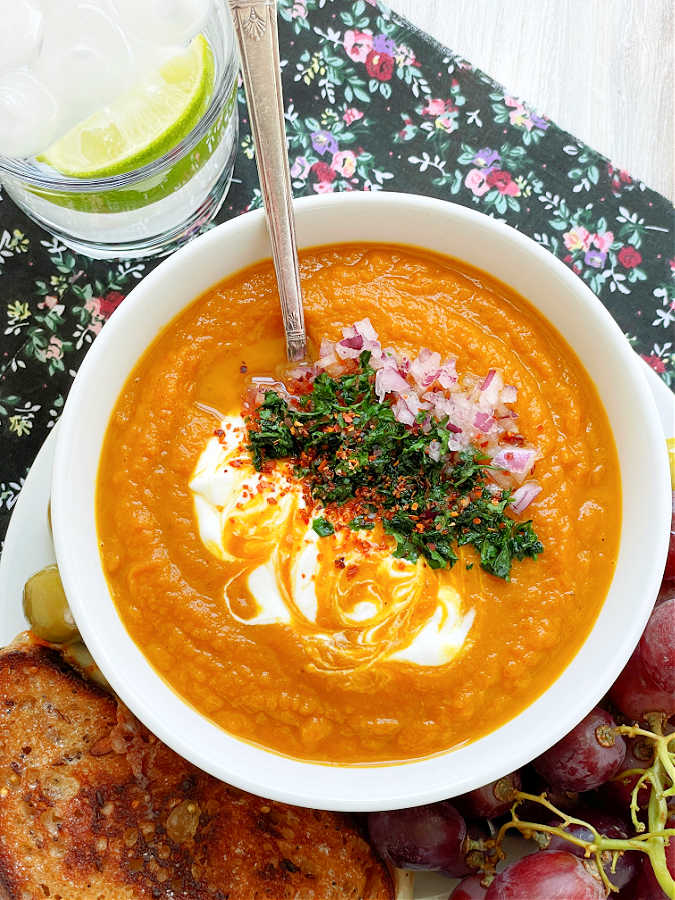 slow cooker pumpkin soup in a bowl garnished with sour cream, red onion and cilantro, plus a grilled cheese sandwich and grapes on the side