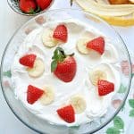 strawberry banana pudding in a glass trifle dish topped with cool whip and fresh sliced berries and bananas