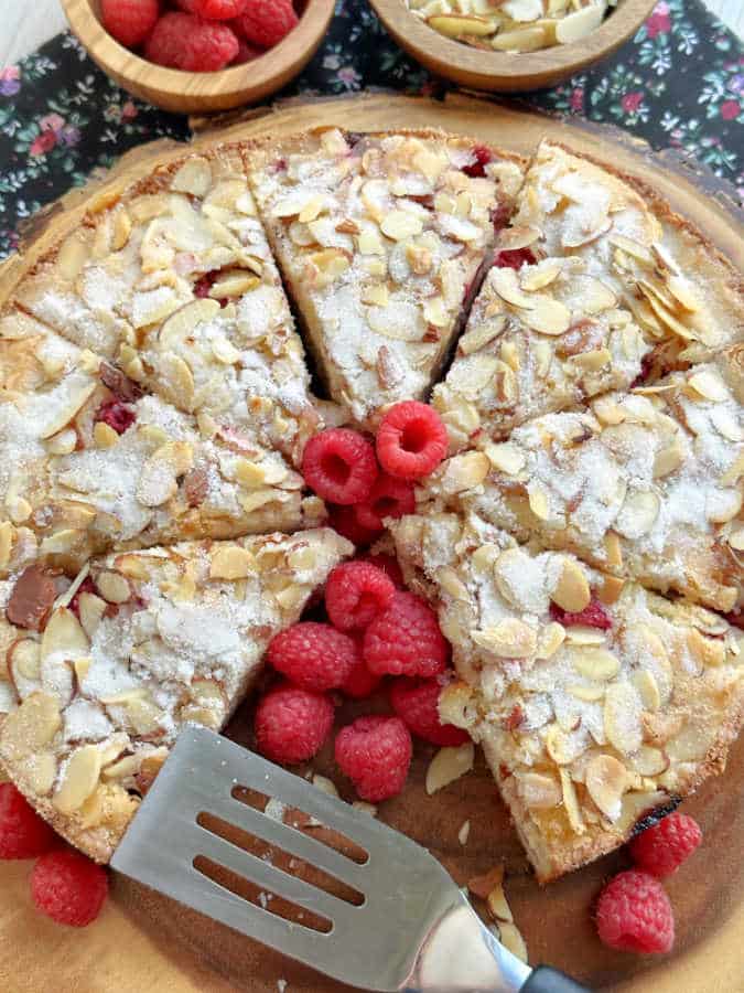 sliced raspberry almond cake on a wooden platter, with fresh raspberries and whipped cream on the side