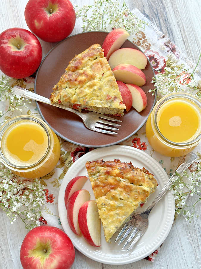An overhead view of two slices of goat cheese quiche plated with apple slices and two glasses of orange juice on the side.