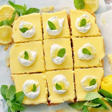 chilled lemon bars, cut into squares. Each square is topped with a small dollop of whipped cream, a fresh lemon slice and a mint leaf