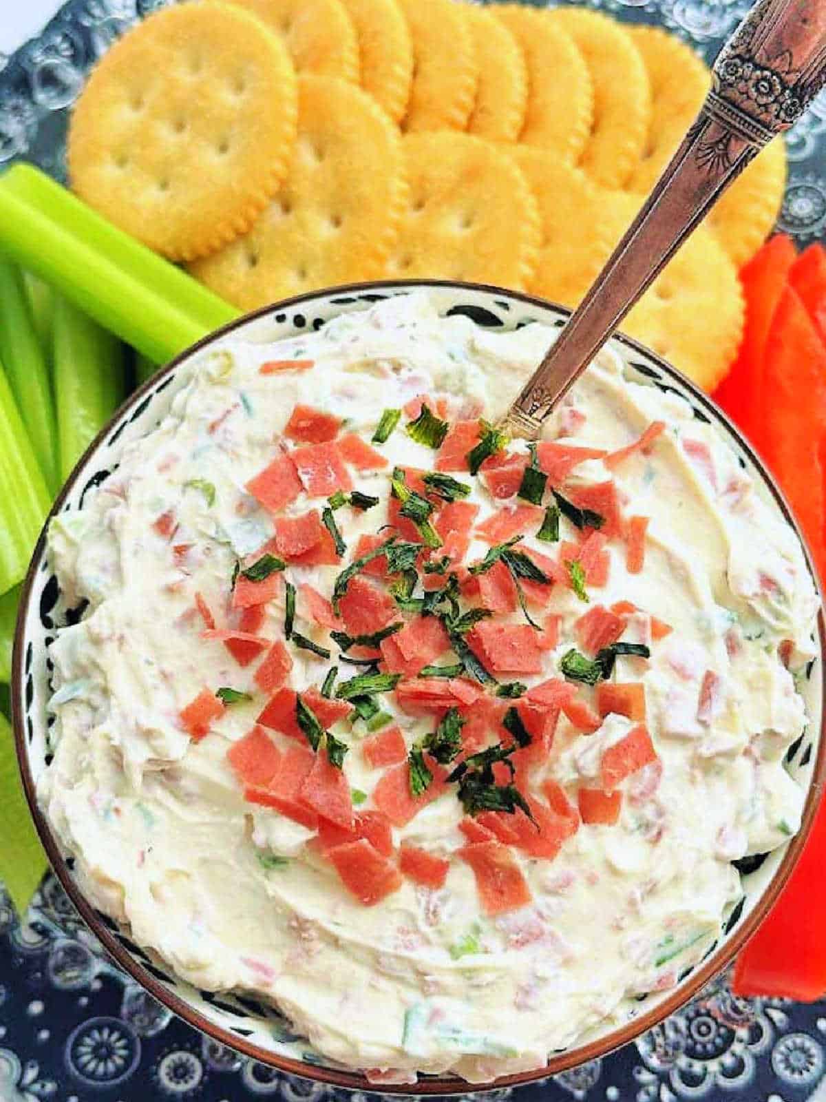 a bowl of dried beef dip surrounded by crackers, celery sticks and red pepper strips