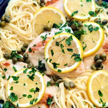 close up of chicken breasts topped with lemon slices and parsley atop a bed of pasta with creamy piccata sauce