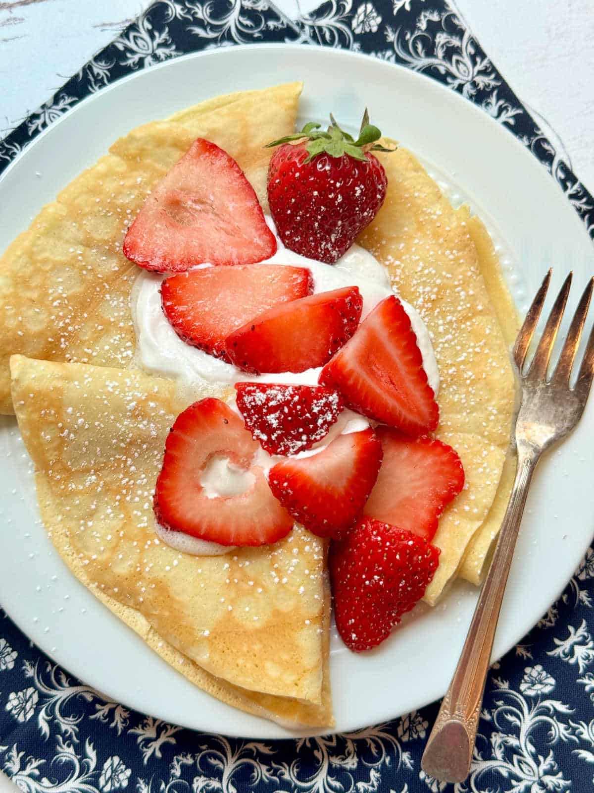 three crepes folded into quarters on a white plate and topped with fresh strawberries and whipped cream