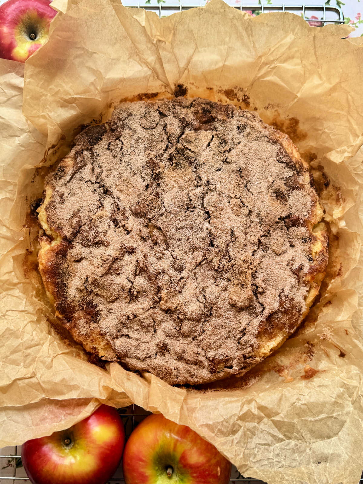 a whole apple cake wrapped in natural colored parchment paper cooling on a wire rack and surrounded by three fresh apples