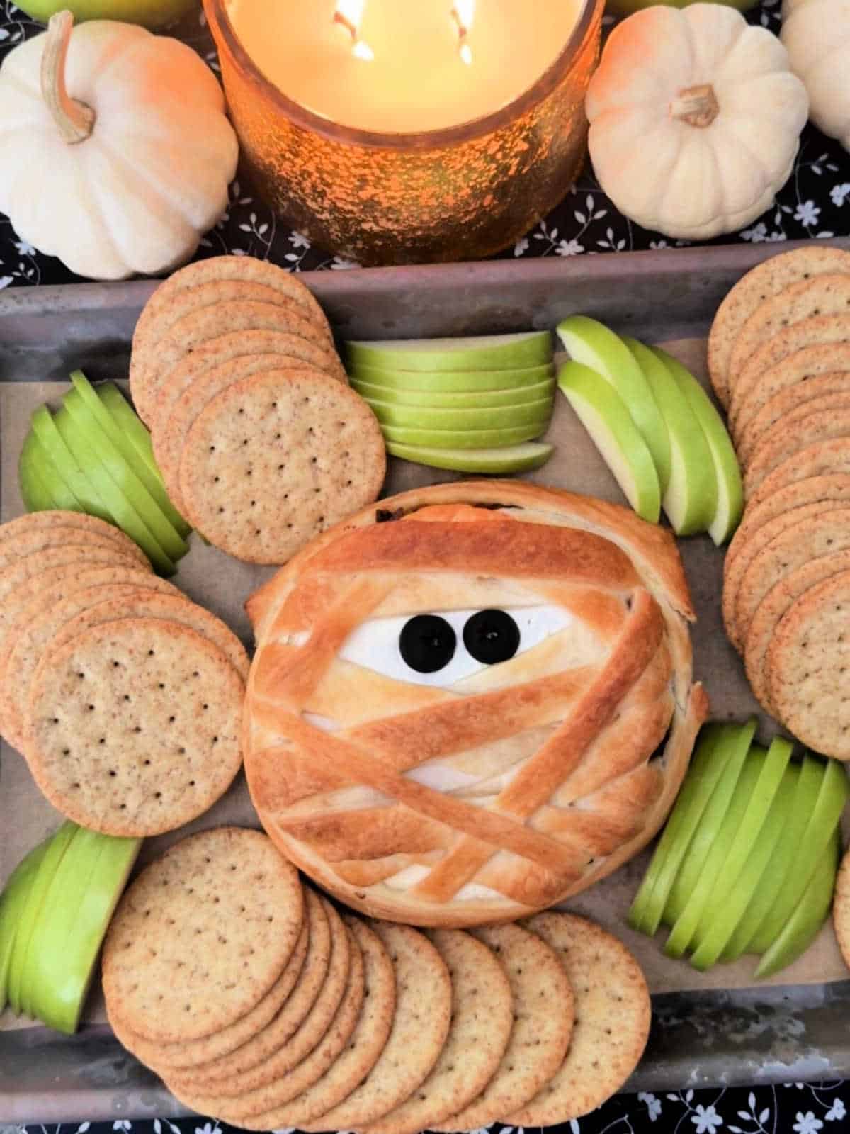 baked mummy brie on the baking sheet surrounded by crackers and green apple slices